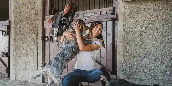 A sitting woman smiles as her dog excitedly puts its paws on her shoulders.